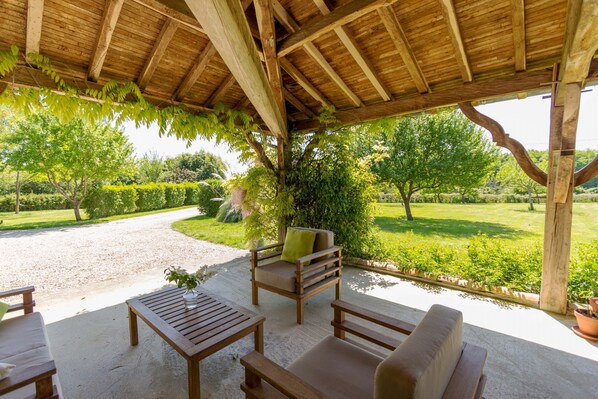 A covered outdoor seating area with wooden furniture and a view of a landscaped garden.