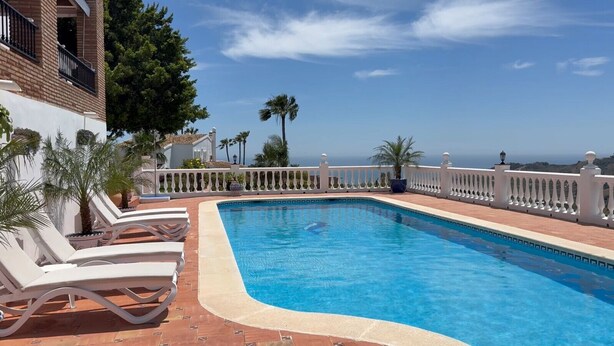 A rectangular swimming pool with a white balustrade, surrounded by lounge chairs and palm trees, overlooking a scenic view.