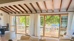 A spacious living room with a large sofa set, a dining table, and a laptop on a desk. Large windows with curtains provide a view of the outdoor patio area.