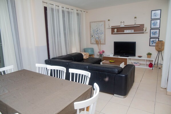 Un salon avec un canapé noir, un téléviseur, une table à manger avec des chaises et une grande fenêtre avec des rideaux.