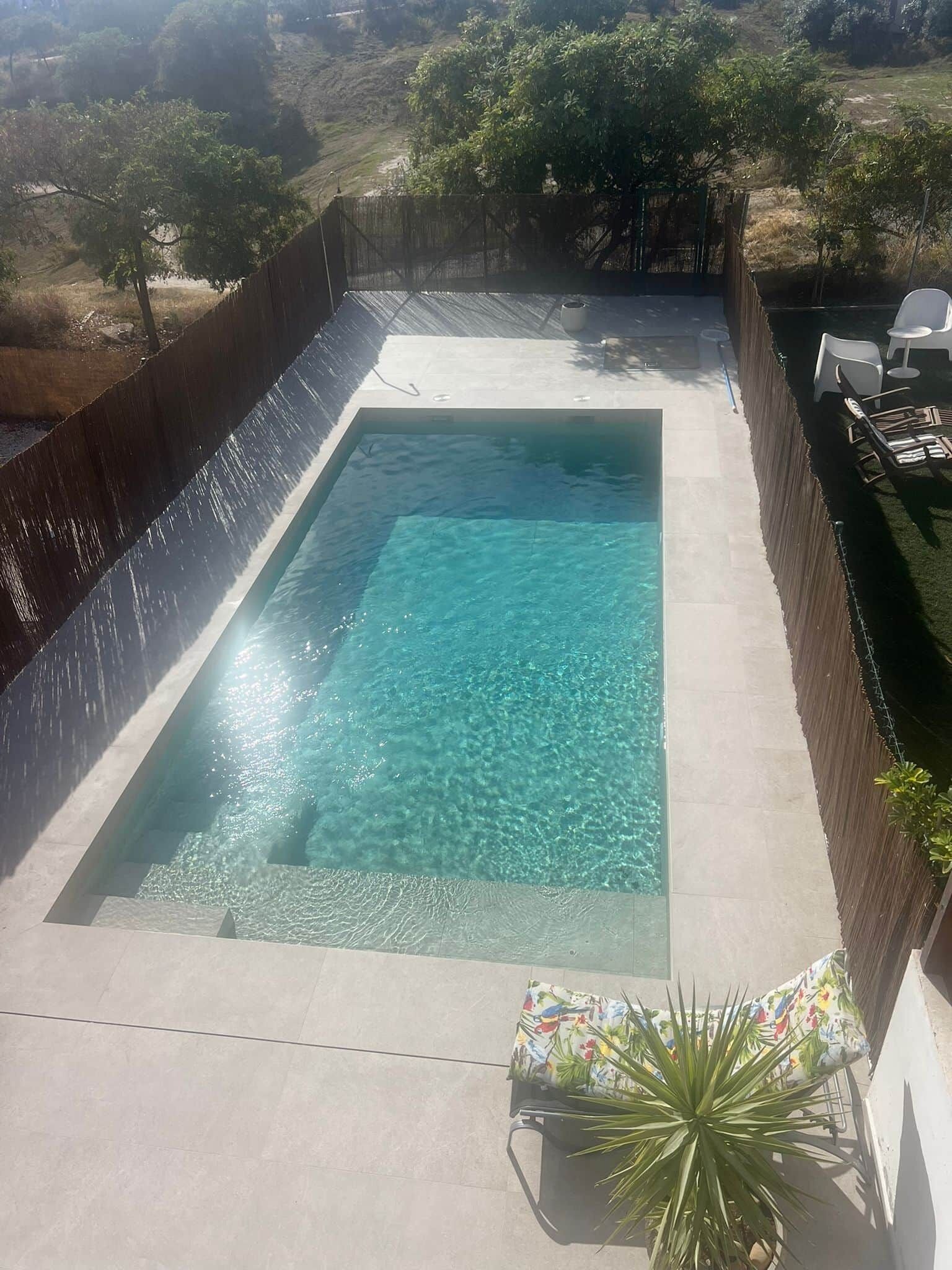Piscina retangular com água cristalina, cercada por uma vedação de madeira e uma área gramada. Há espreguiçadeiras brancas e uma mesa pequena de um lado.