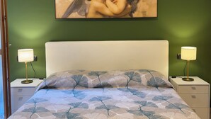 Ein Schlafzimmer mit grüner Wand, einem Cherubengemälde, einem weißen Bett, zwei Nachttischlampen und einer gemusterten Bettdecke.