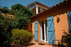 A house with a tiled roof, a door with blue shutters, and a small garden with bushes and a tree.