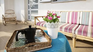 Una bandeja de mimbre con una cafetera y tazas sobre un mantel azul, una silla de mimbre, un sofá a rayas y un jarrón blanco con flores.