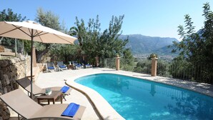 Une piscine entourée de chaises longues et d’un parasol, bordée de murs de pierre et d’arbres, avec une vue sur les montagnes en arrière-plan.