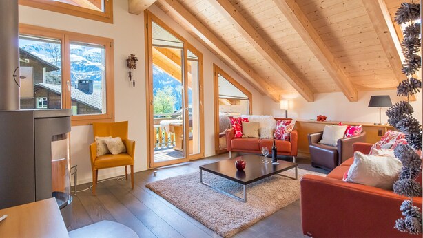 A cozy living room with a fireplace, wooden beams, and a view of mountains through large windows. - Duplex Penthouse in Chalet Luna (Wengen)