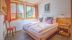 A bedroom with a wooden bed, a chair, a nightstand, and a window with curtains. - Duplex Penthouse in Chalet Luna (Wengen)
