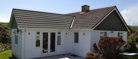 A single-story house with a grey tiled roof, white exterior walls, and a small front yard with a lawn and shrubbery.