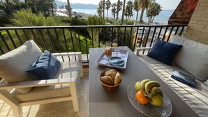 A balcony with a sofa, a table with a tray of food and drinks, and a view of the beach and palm trees.