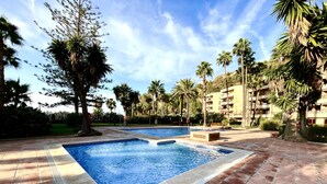 A swimming pool with a tiled deck, surrounded by palm trees and a multi-story building.