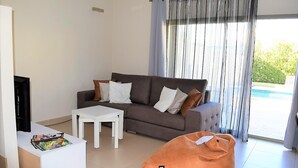Un salon moderne avec un canapé gris, une table basse blanche et un fauteuil orange. Un téléviseur est fixé au mur, et une fenêtre avec des rideaux légers est visible.