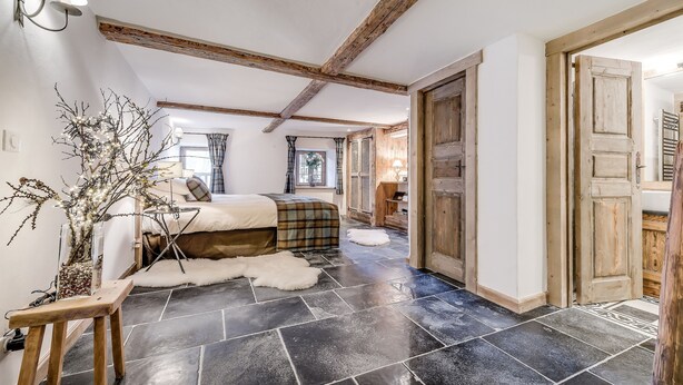 Une chambre à coucher avec un lit, un banc en bois, un vase avec des branches et un sol en pierre.