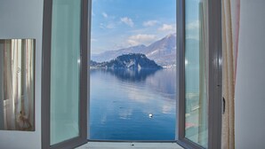 A room with a large window offering a view of a calm lake and distant mountains. - Apartment with balcony, direct beach access (Varenna)