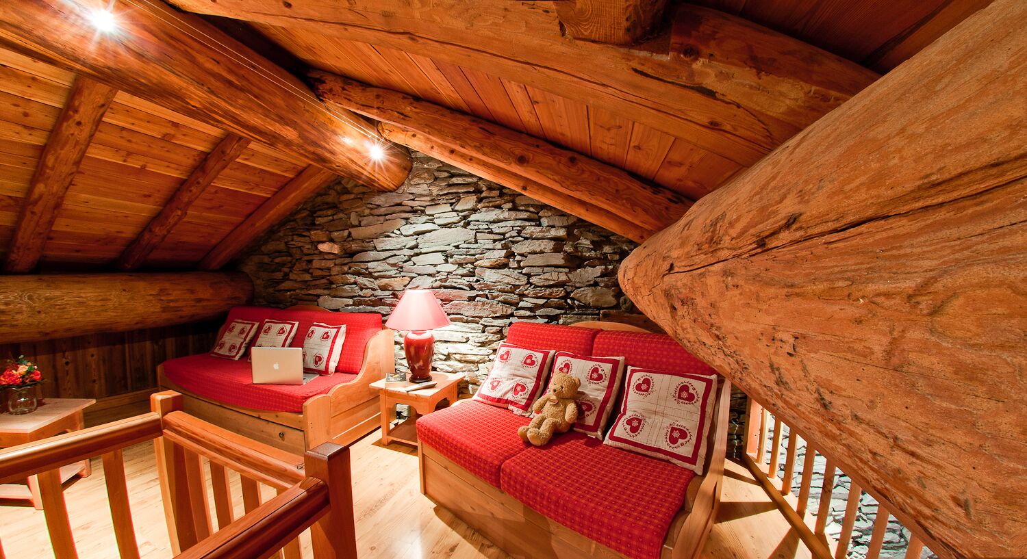 A cozy attic bedroom with a stone wall, wooden beams, a red sofa, and a wooden chair.