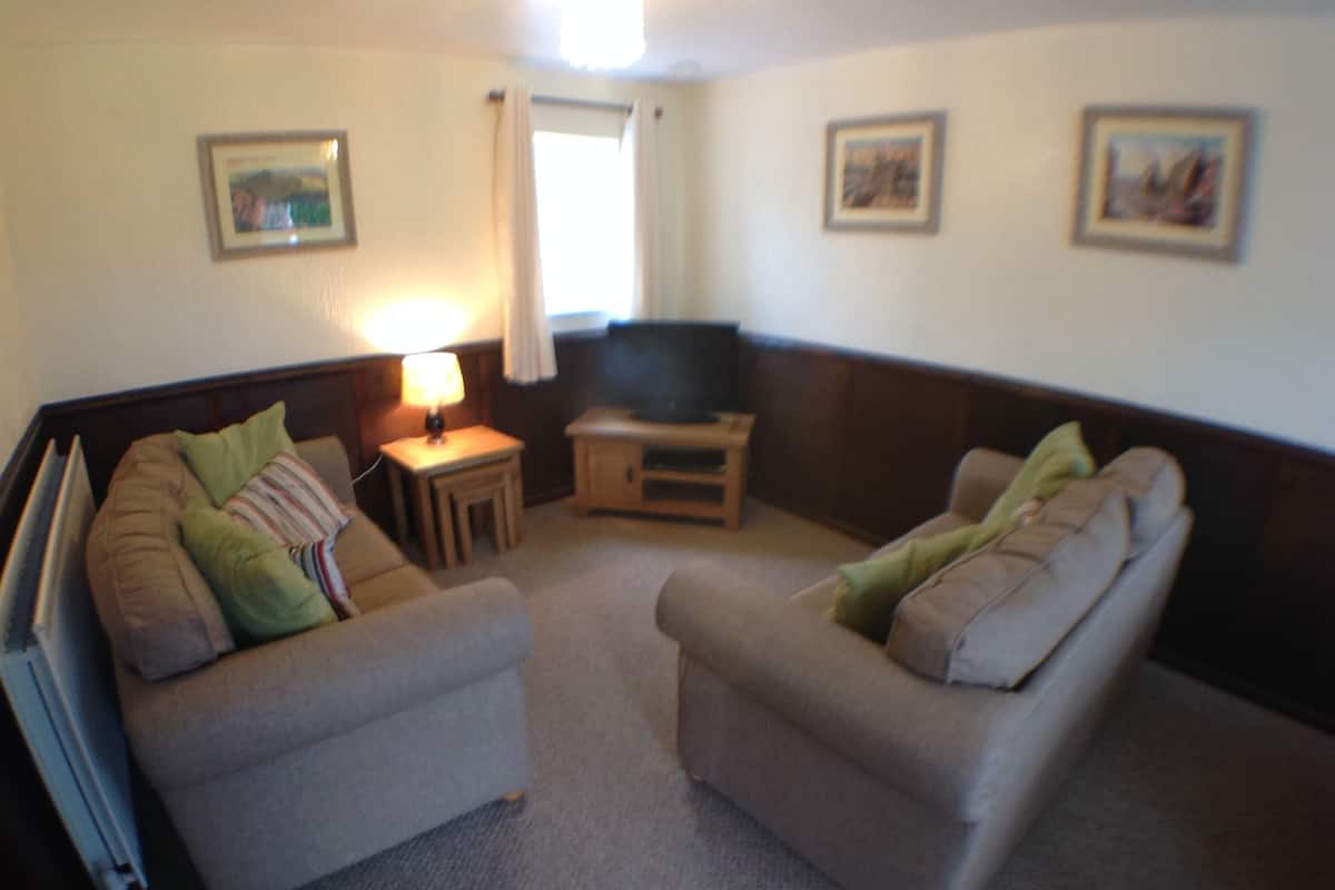 A living room with two grey sofas, a small table with a lamp, a television, and framed pictures on the walls.