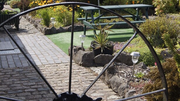 A garden with a large metal wheel, stone pathway, potted plants, and a green bench. - Birchenfields Family Friendly Cottages, Play Barn for all Ages and Summer Hous (Stoke-on-Trent)