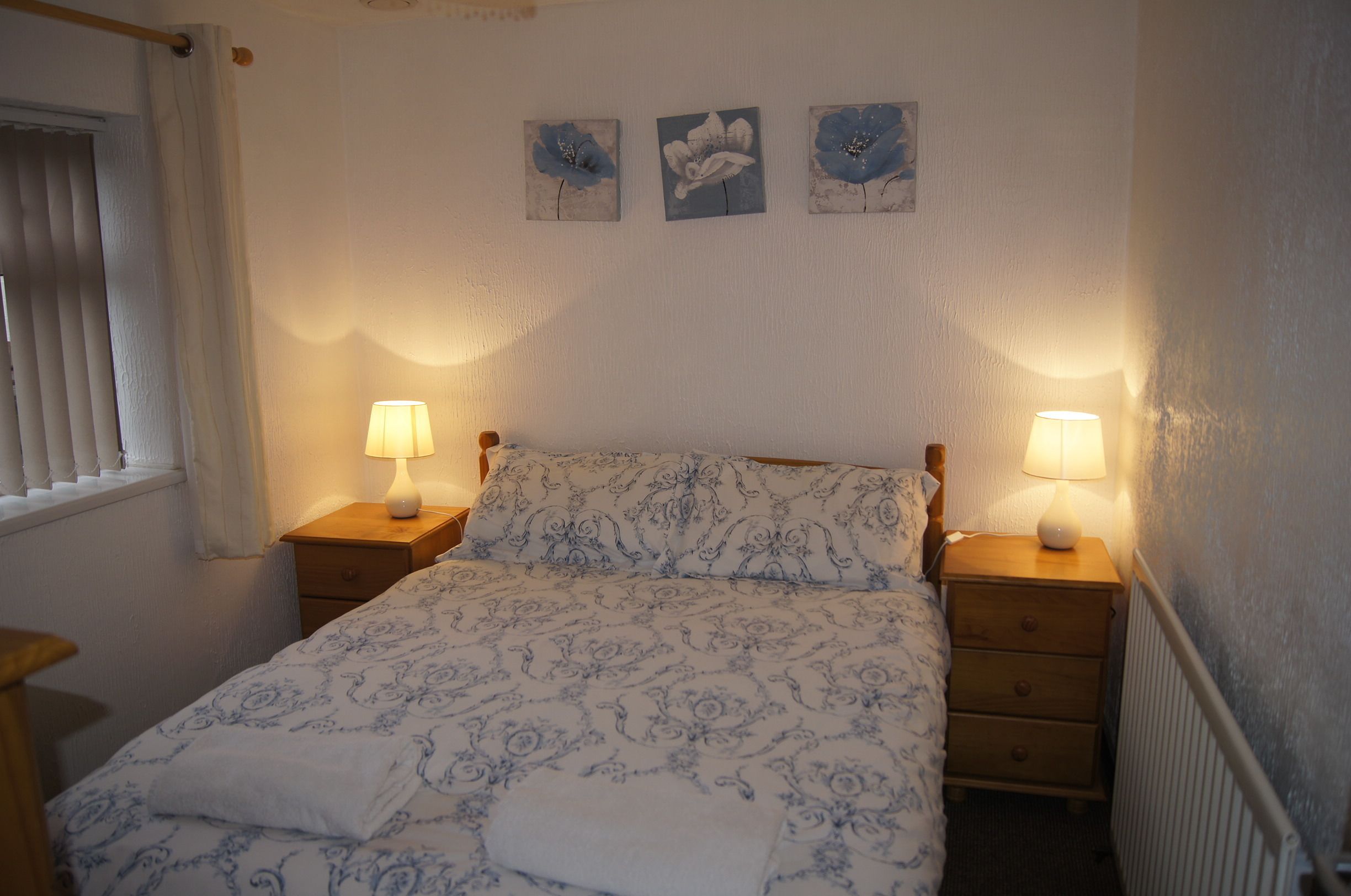 A bedroom with a bed, two bedside tables with lamps, a window with blinds, and three wall-mounted paintings.