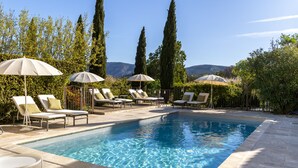 A rectangular swimming pool with clear blue water, surrounded by lounge chairs and umbrellas, set against a backdrop of tall trees and distant mountains. - Apartment/ flat - Lagnes-L'Isle-sur-la-Sorgue (lAGNES)