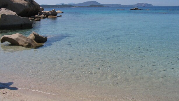 Een strand met helder water, grote rotsen en bergen in de verte.