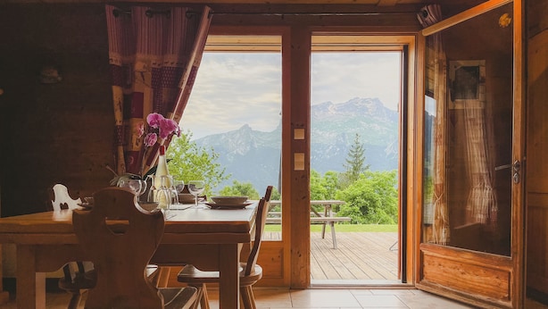 A wooden dining table with chairs, a vase with flowers, and a view of mountains through an open door.