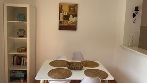 A dining area with a white table, four white chairs, and woven placemats. A bookshelf with books and decorative items is to the left. - Villa in Gosici, Radovici. Private Pool, BBQ, peaceful location, stunning views. (Gošići)