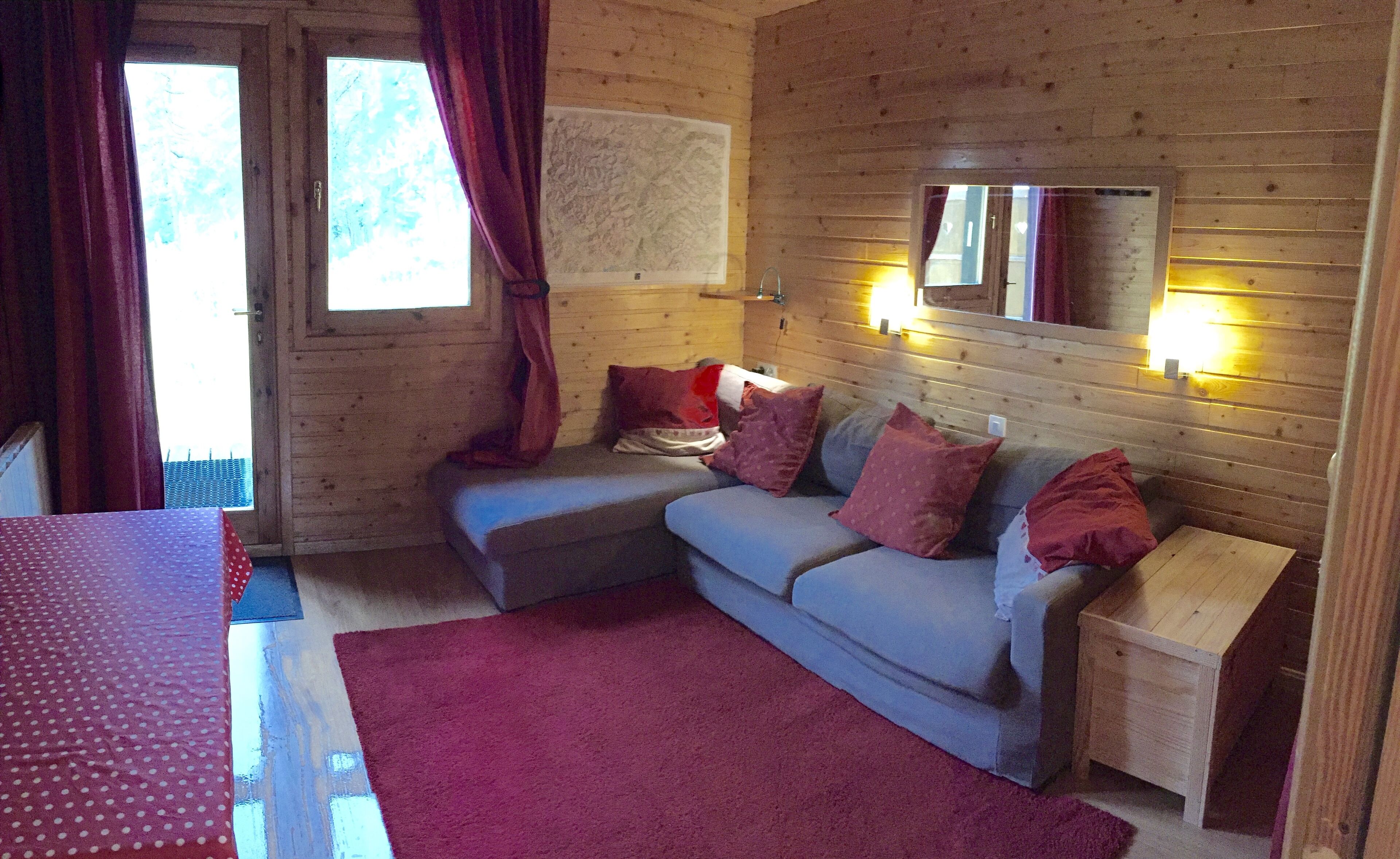 A cozy room with a wooden interior, a grey sofa with red cushions, a wooden nightstand, and a red rug.