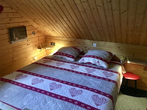 A cozy wooden cabin bedroom with a bed, pillows, a nightstand, and a red chair.