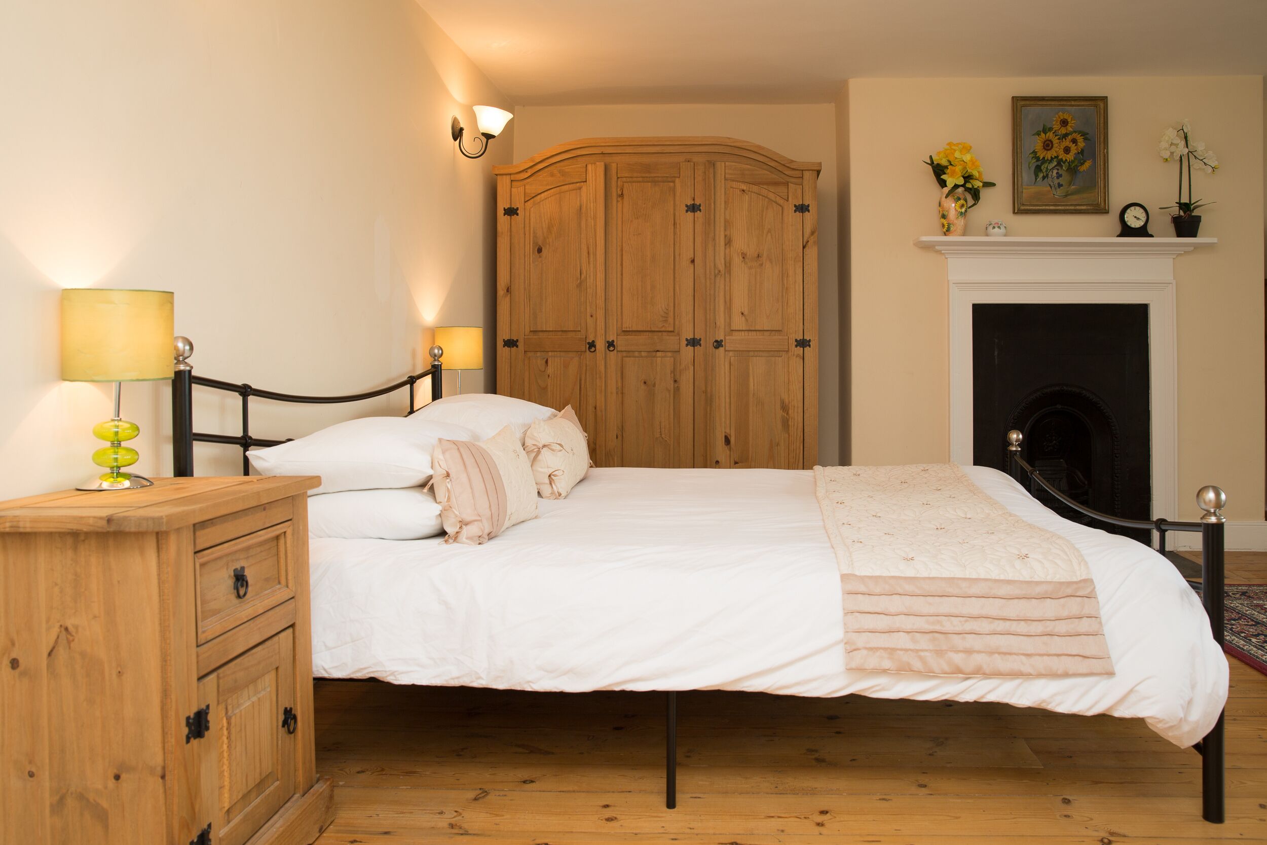 A bedroom with a wooden wardrobe, a bed with white bedding, a nightstand with a lamp, and a fireplace.
