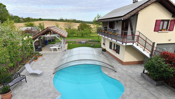Een zwembad met een overdekt gedeelte, een terras met zitplaatsen, en een huis met een balkon.
