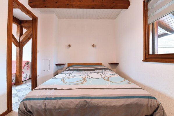 A bedroom with a wooden headboard, a bed with striped bedding, a window with blinds, and a door leading to another room.