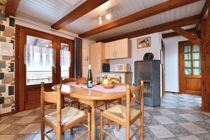 A dining area with a wooden table and chairs, a fireplace, a kitchen area with cabinets, and a window with curtains. - Country Cottage / Gite - Hirtzbach (Hirtzbach)