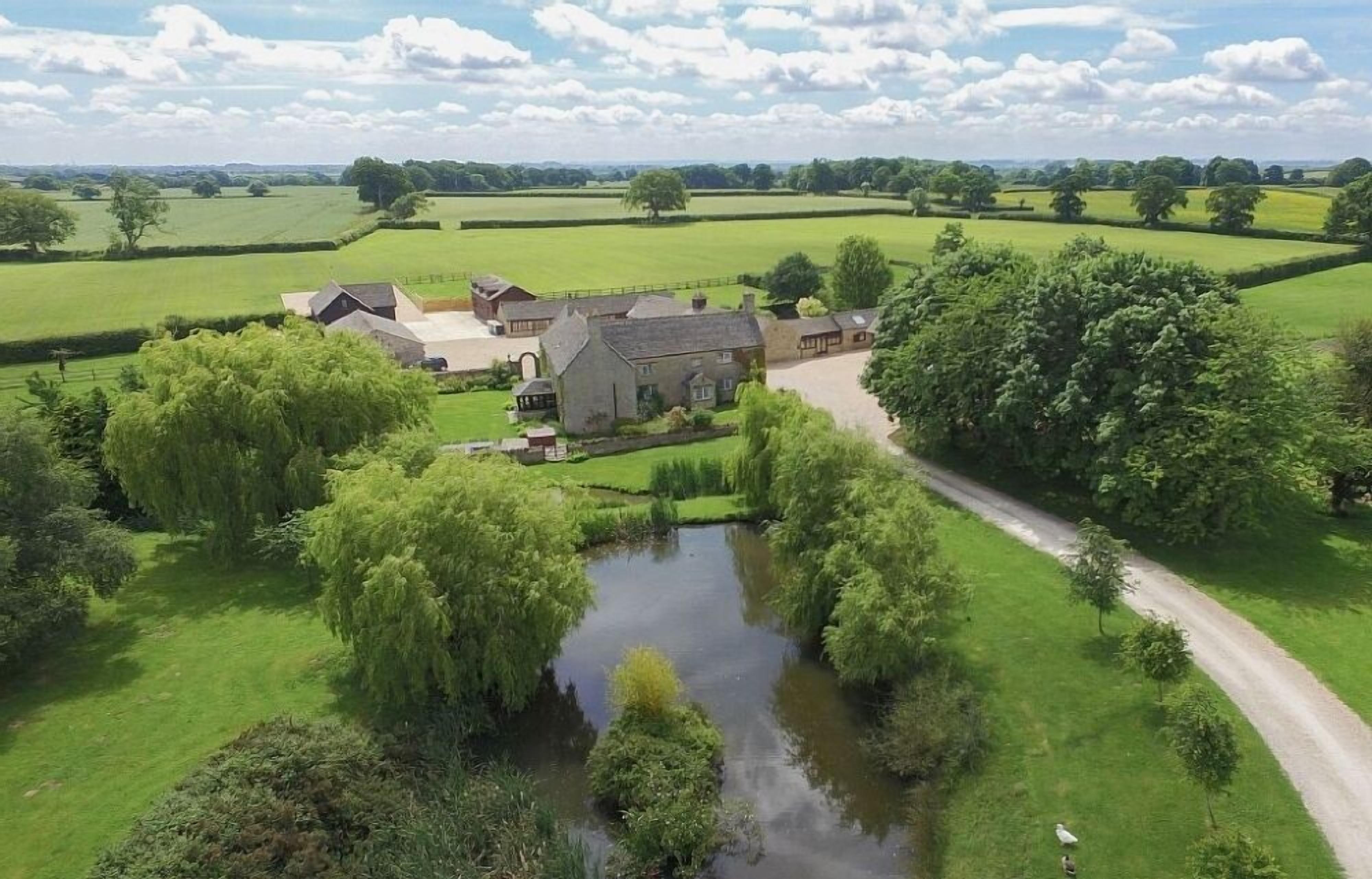 A rural estate with a pond, a pathway, and a large house surrounded by fields and trees.