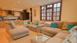 A living room with a sofa set, a coffee table with magazines, and a kitchen in the background. - The Cotswold Manor Byre, Exclusive Hot Tub, Games Barn, 70 acres of Parkland (Bampton)