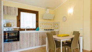 A compact kitchen with a built-in oven, a gas stove, and a window with curtains. There is a dining table with chairs and a yellow tray. - Villaggio Perlacea - PERLACEA 34 (Golfo Aranci)