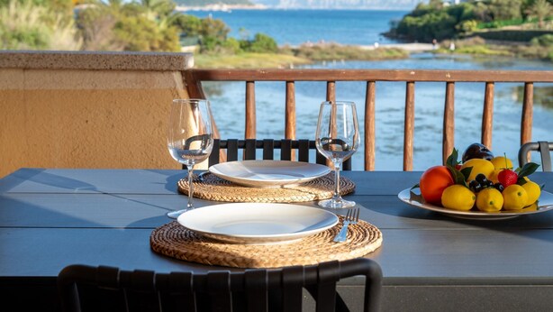 A table set with plates, glasses, and a fruit plate, overlooking a coastal view with mountains. - Villaggio Perlacea - PERLACEA 34 (Golfo Aranci)
