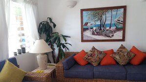 Ein Wohnzimmer mit einem blauen Sofa, orangefarbenen Kissen, einem Korbtisch, einer Lampe und einem Gemälde mit Strandmotiv.