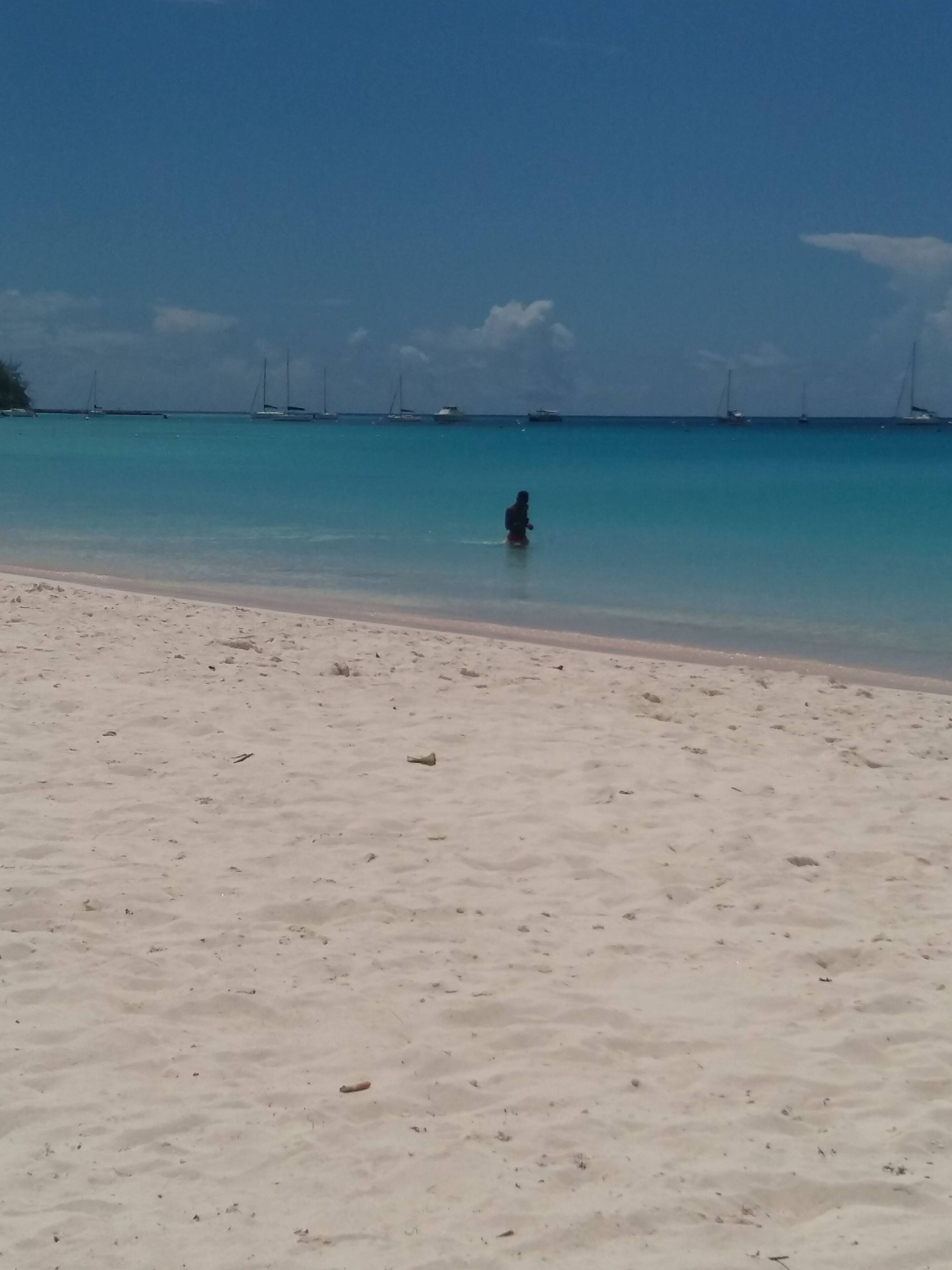 A beach with clear turquoise water and a few sailboats in the distance.