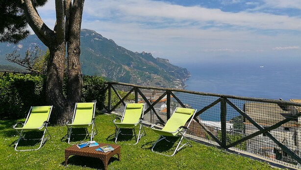 A view of the sea with a mountain in the background, two green lounge chairs, and a wicker table with magazines.