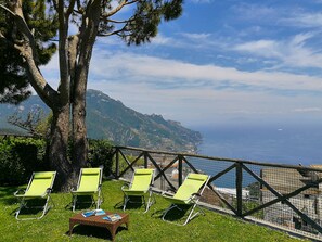 Une vue sur la mer, avec une montagne en arrière-plan, deux fauteuils de jardin verts et une table en osier sur laquelle sont posés des magazines.