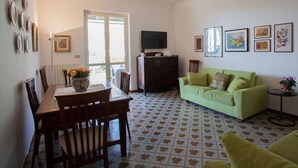 A living room with a green sofa, a dining table with chairs, a TV on a stand, and a patterned floor.