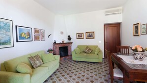 A living room with two green sofas, a fireplace, a dining table with chairs, and a tiled floor.