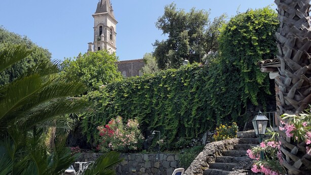 Zona de piscina con una escalera de piedra, abundante vegetación y una torre de iglesia al fondo.