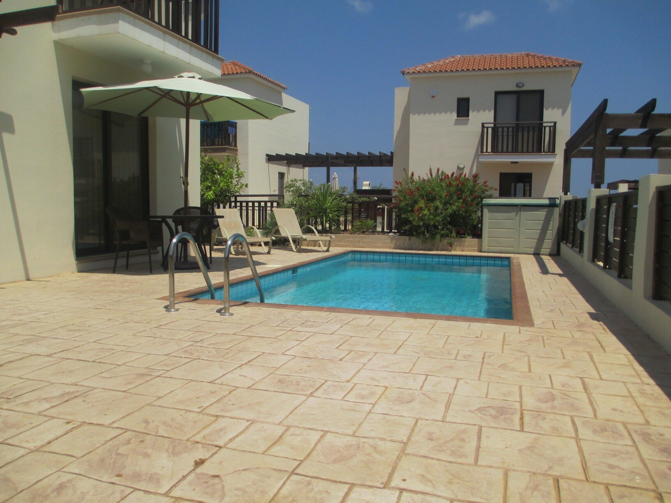 A swimming pool with lounge chairs and a table, surrounded by buildings with balconies and a tiled floor.