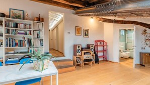 A spacious living room with wooden flooring, a white table, and a bookshelf filled with books. There is a staircase leading to another room, and a red chair in the corner. - Gabry's Rooftop Terrace Home with  breathtaking Views of Lucca & Tuscany (Lucca)