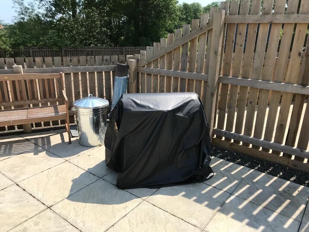 Een overdekte barbecue en een vuilnisbak op een terras met een houten omheining op de achtergrond.
