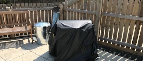 Een overdekte barbecue en een vuilnisbak op een terras met een houten omheining op de achtergrond.
