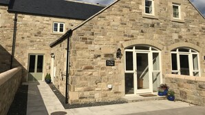 Un bâtiment en pierre avec un toit noir, une enseigne indiquant « Pyg Pen Cottage », et un petit porche agrémenté de plantes en pot.