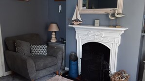 A living room with a grey sofa, a small table with a lamp, a fireplace, and a blue barrel. - Amber Cottage 3 Bed , In Snettisham, North Norfolk  dogs welcome (Snettisham)