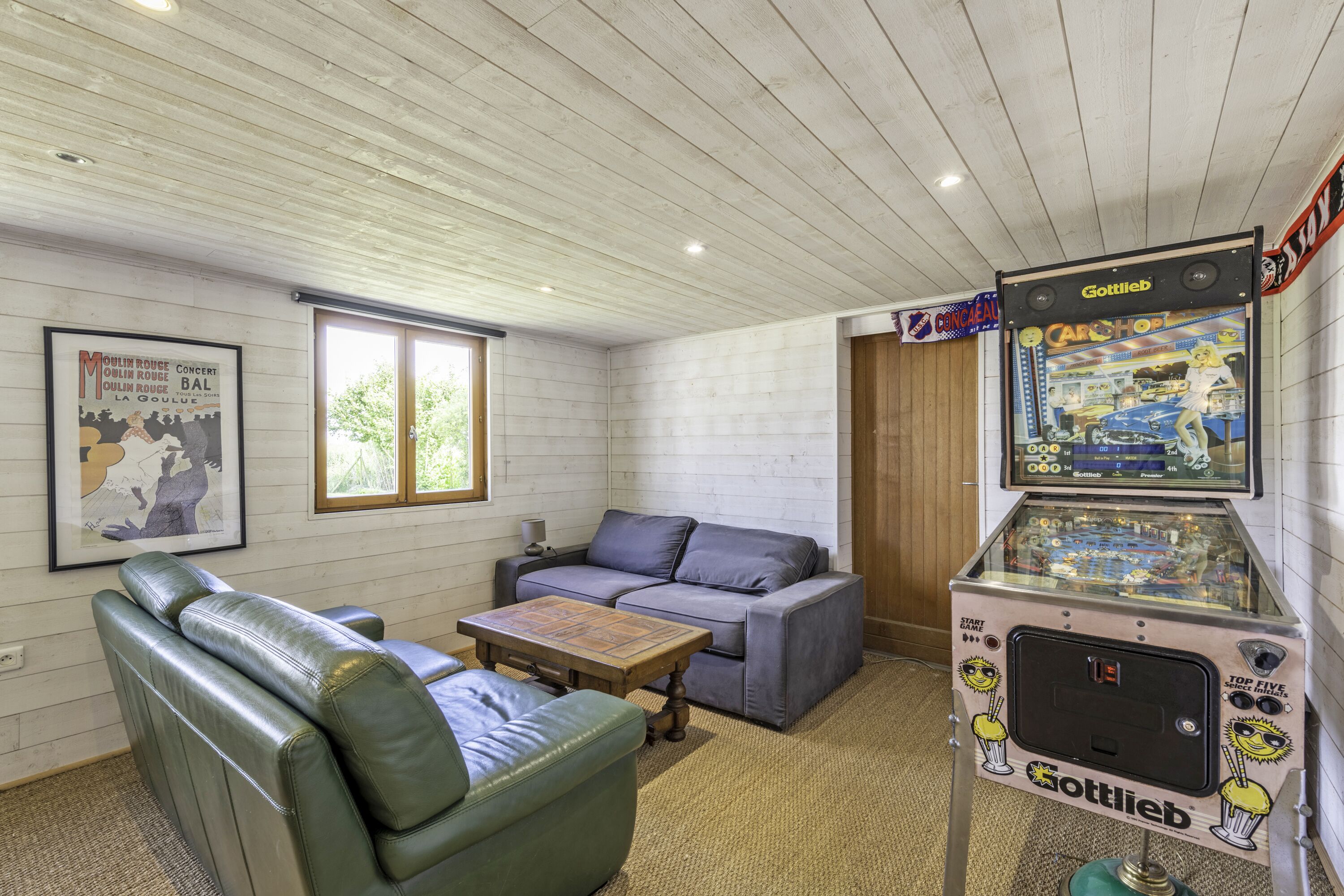A room with a wooden ceiling, a grey sofa, a green armchair, a small wooden table, and a Gottlieb arcade machine.