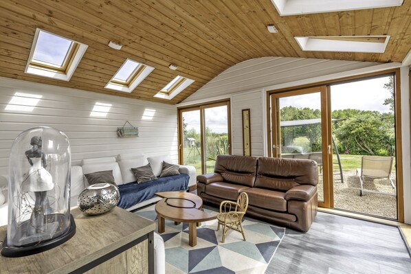 A modern living room with a wooden ceiling, large windows, a brown leather sofa, a white sofa, and a glass coffee table. - A beachfront cottage (Cléder)